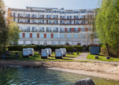 Blick auf die Hotelrückseite mit Balkonfassade, bepflanzten Wegen, Strandkörben und großen Ziersträuchern – barrierearmer Zugang zu Gartenflächen möglich.