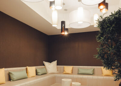 Nische mit Sitzpolstern in Beige und Grün, Holztisch und Pflanzen – ruhiger, barrierefreier Warte- oder Gesprächsbereich.