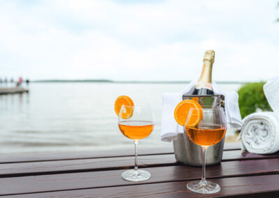Eine Flasche Champagner im Kühler, daneben zwei Gläser mit orangefarbenem Getränk, Hintergrund See und Steg.