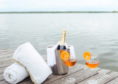 Eine Flasche Champagner im Kühler, daneben zwei Gläser mit orangefarbenem Getränk, Hintergrund See und Steg.