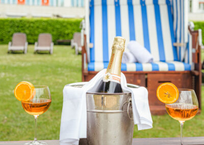 Eine Flasche Champagner im Kühler, daneben zwei Gläser mit orangefarbenem Getränk, Hintergrund Strandkorb.