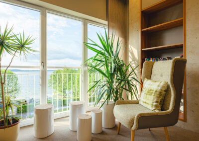Gemütliche Leseecke mit Polstersessel, Regal mit Büchern und Dekoobjekten, stilvolle Raumbeleuchtung, Fenster mit Blick auf Natur, geeignet als ruhiger Rückzugsort.